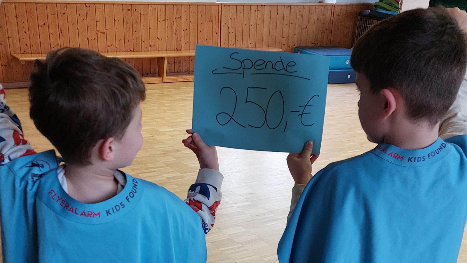 Zwei Kinder in blauen Umhängen halten ein Plakat mit 'Spende 250€' in einem Raum mit Holzboden.