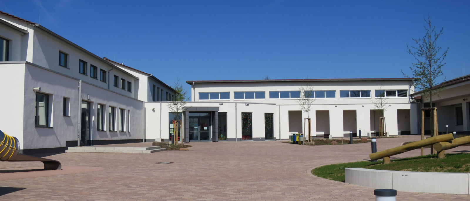 Moderner Schulhof mit hellen Gebäuden, Pflastersteinen und einem blauen Himmel.