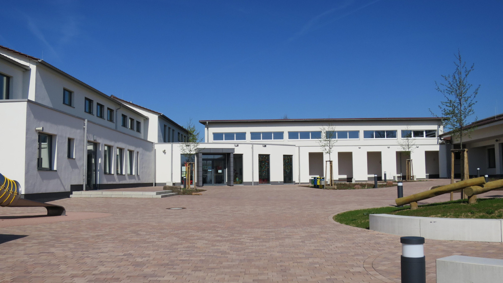 Moderner Schulhof mit hellen Gebäuden, Pflastersteinen und einem blauen Himmel.