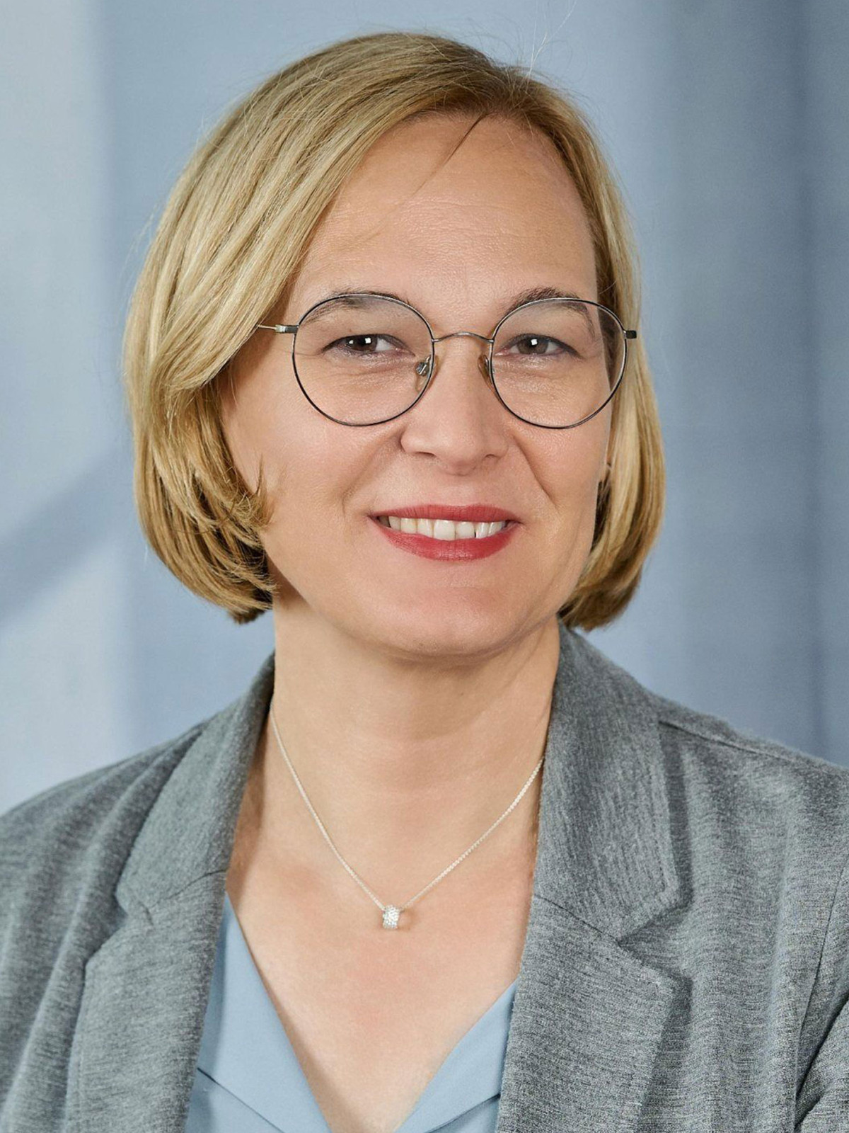 Porträt einer lächelnden Frau mit blonden Haaren und Brille im grauen Blazer.
