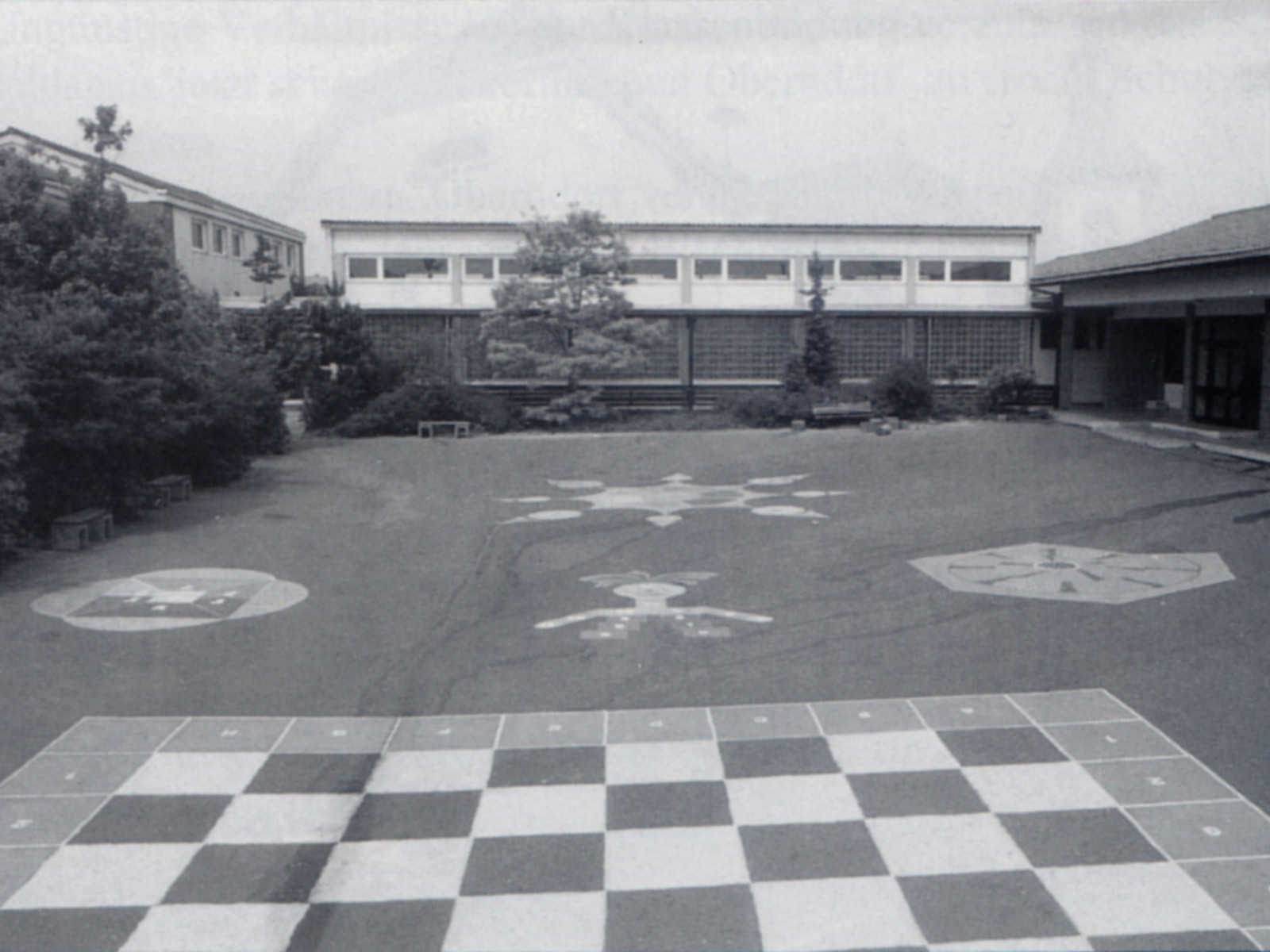 Schulhof mit bemaltem Schachbrettmuster auf Asphalt, umgeben von Schulgebäuden.