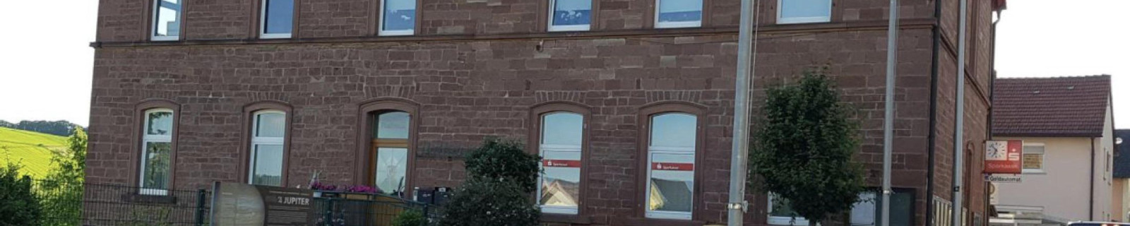 Ein zweistöckiges Steingebäude mit roten Ziegeln und mehreren Fenstern, umgeben von Pflanzen.