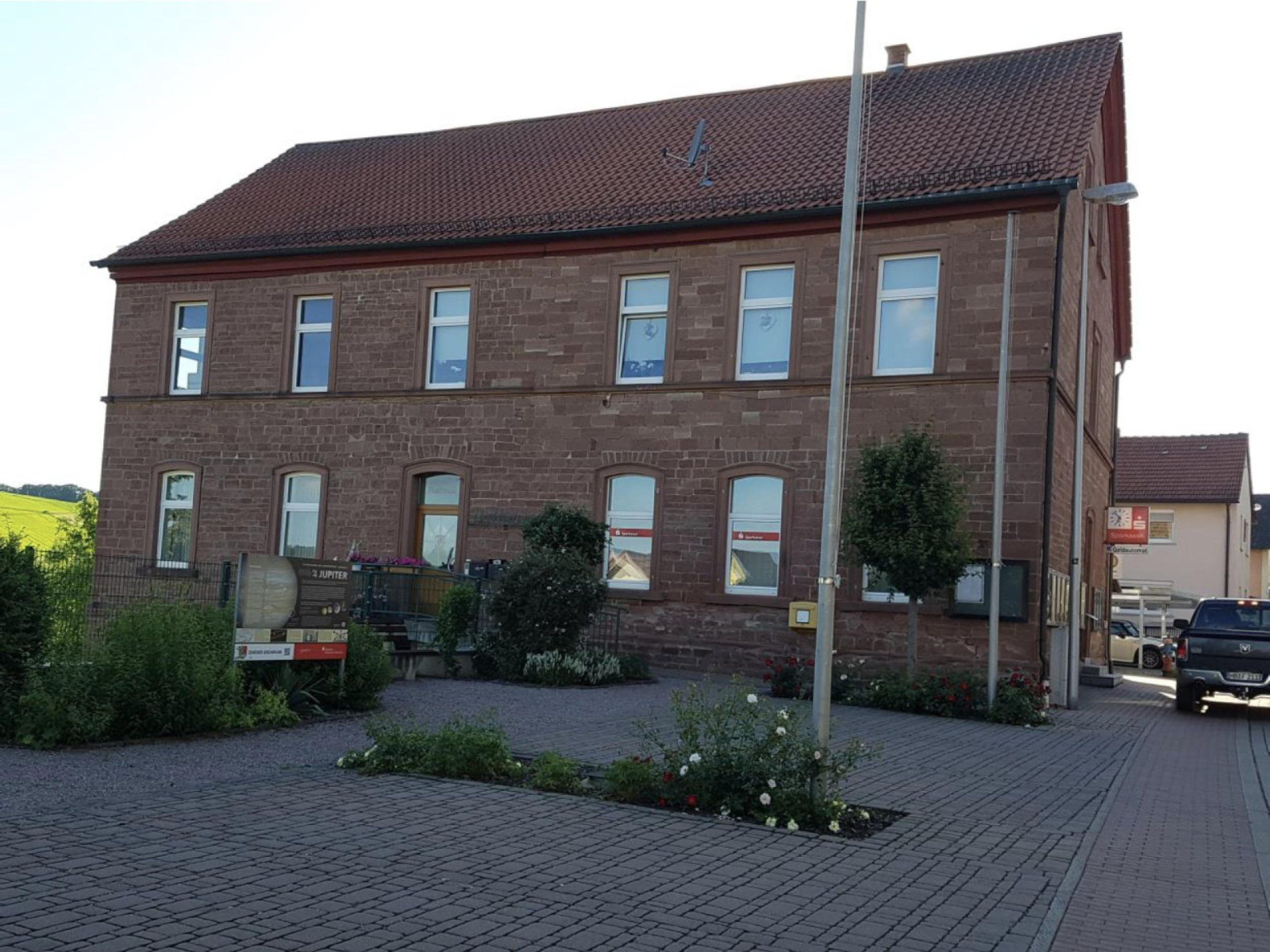 Zweistöckiges historisches Gebäude mit rotem Dach, umgeben von Pflanzen, mit einem Auto rechts.