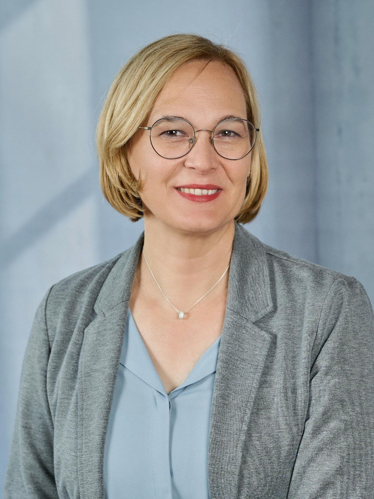 Frau mit Brille, blondem Haar, in grauem Blazer und hellblauer Bluse lächelt.