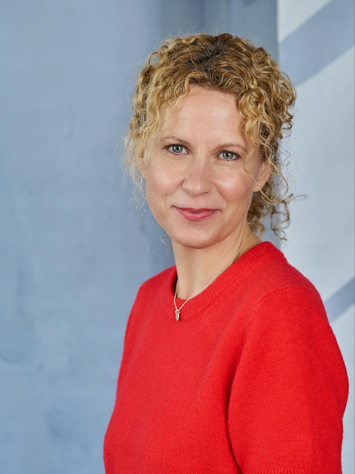 Lächelnde Frau mit lockigem, blondem Haar in rotem Pullover vor blauem Hintergrund.