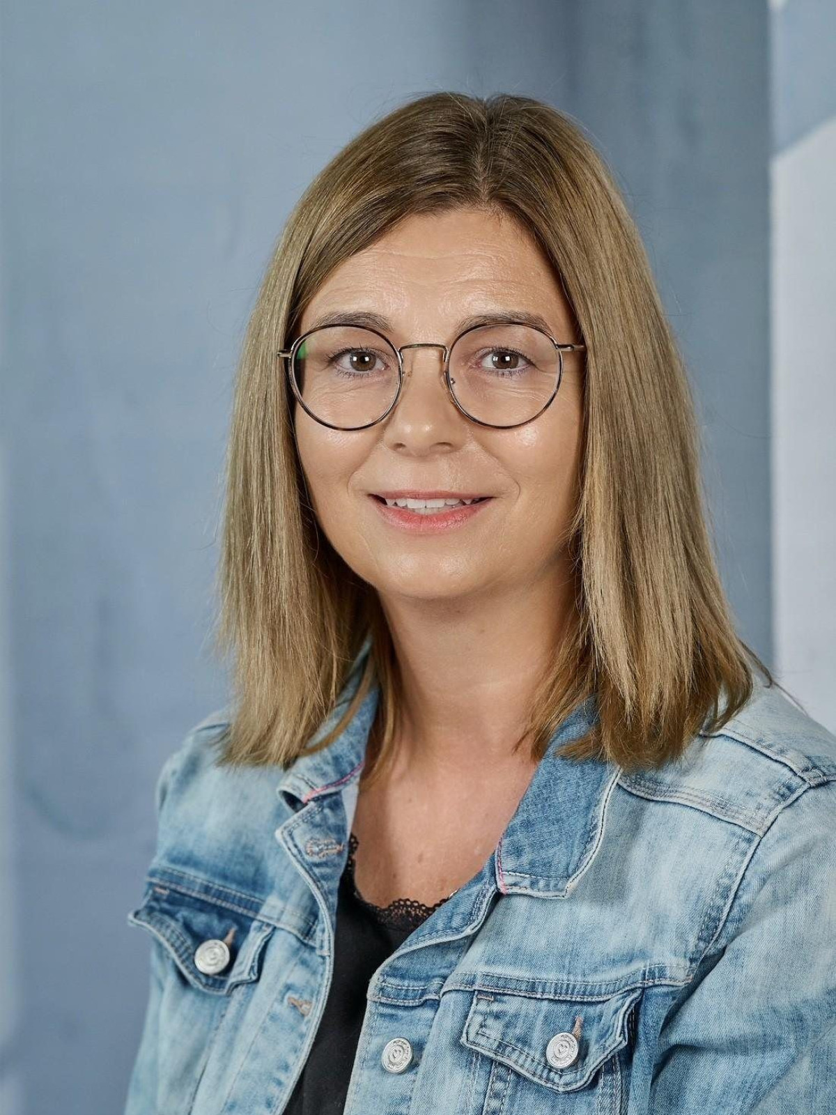 Frau mit Brille und Jeansjacke lächelt vor neutralem Hintergrund.