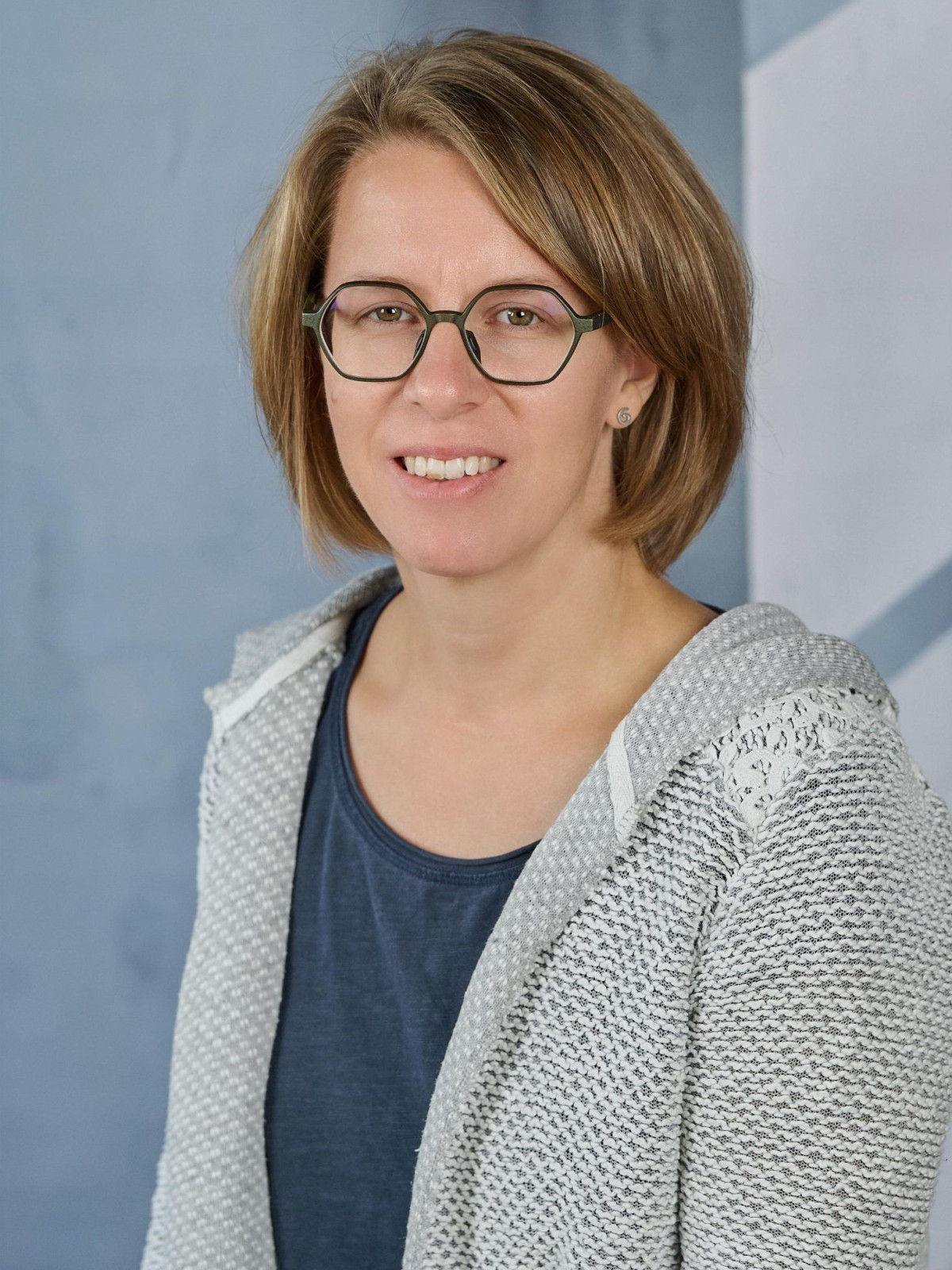 Frau mit Brille und schulterlangem Haar, grauer Pullover, blauer Hintergrund.