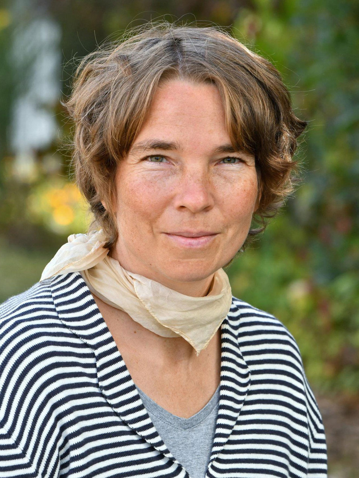Frau mit kurzem, braunem Haar und gestreiftem Pullover und Halstuch, Hintergrund unscharf.