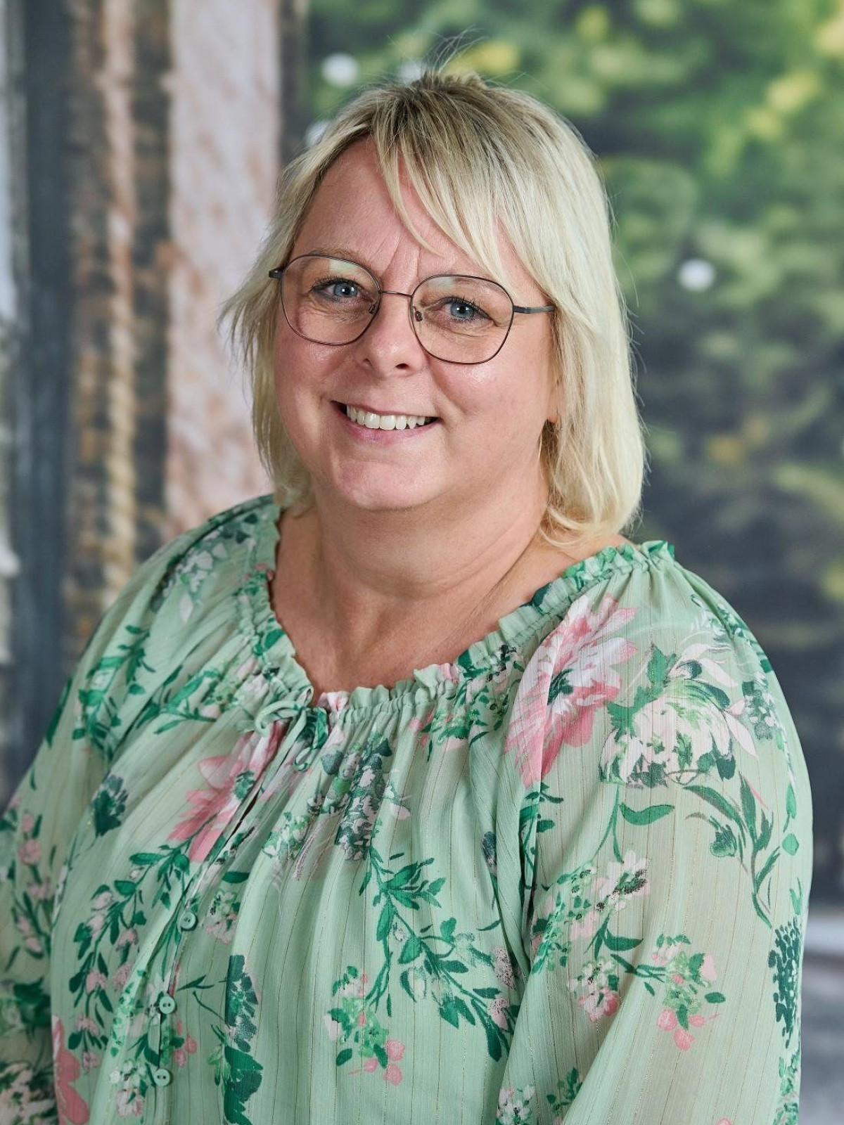 Lächelnde Frau mit Brille und floralem Oberteil vor einem grünen Hintergrund.
