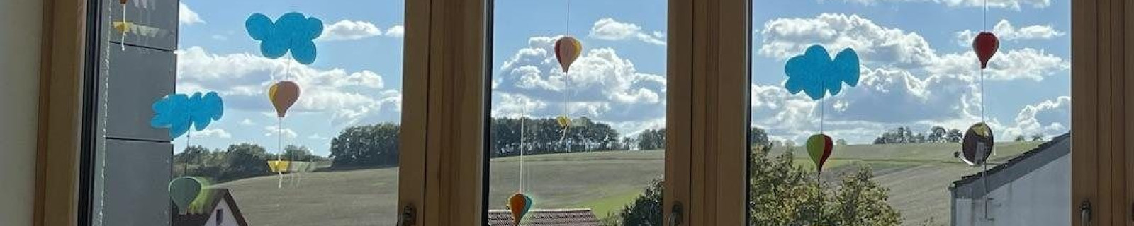Blick durch Fenster mit sonnigem Himmel, Wolken und Landschaft. Bunte Dekorationen hängen an den Scheiben.