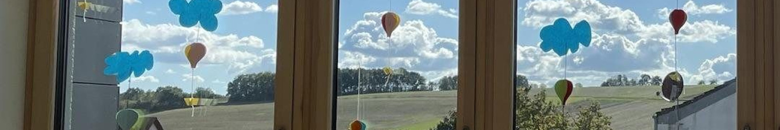 Blick durch Fenster mit sonnigem Himmel, Wolken und Landschaft. Bunte Dekorationen hängen an den Scheiben.