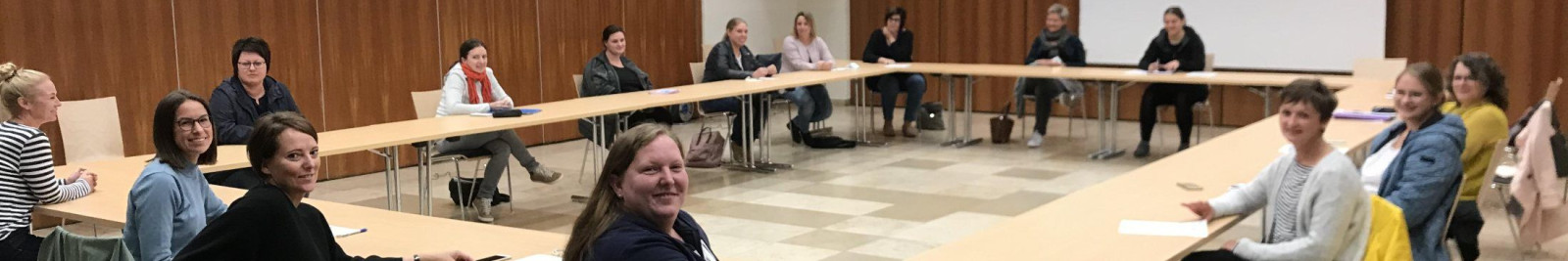 Gruppe von Personen sitzt in einem großen Raum an einem U-förmigen Tisch.