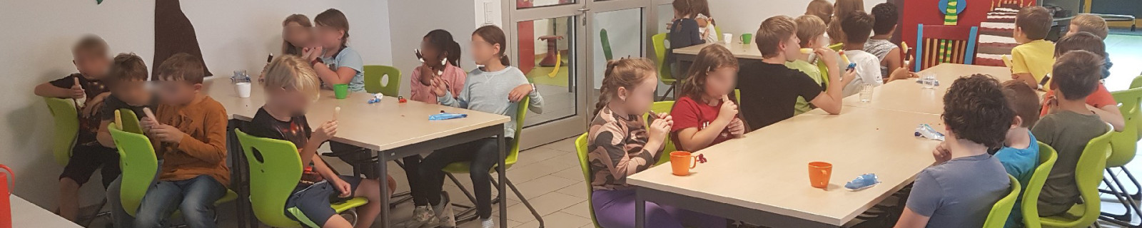 Kinder essen gemeinsam an Tischen in einem hellen Raum mit Baum an der Wand.