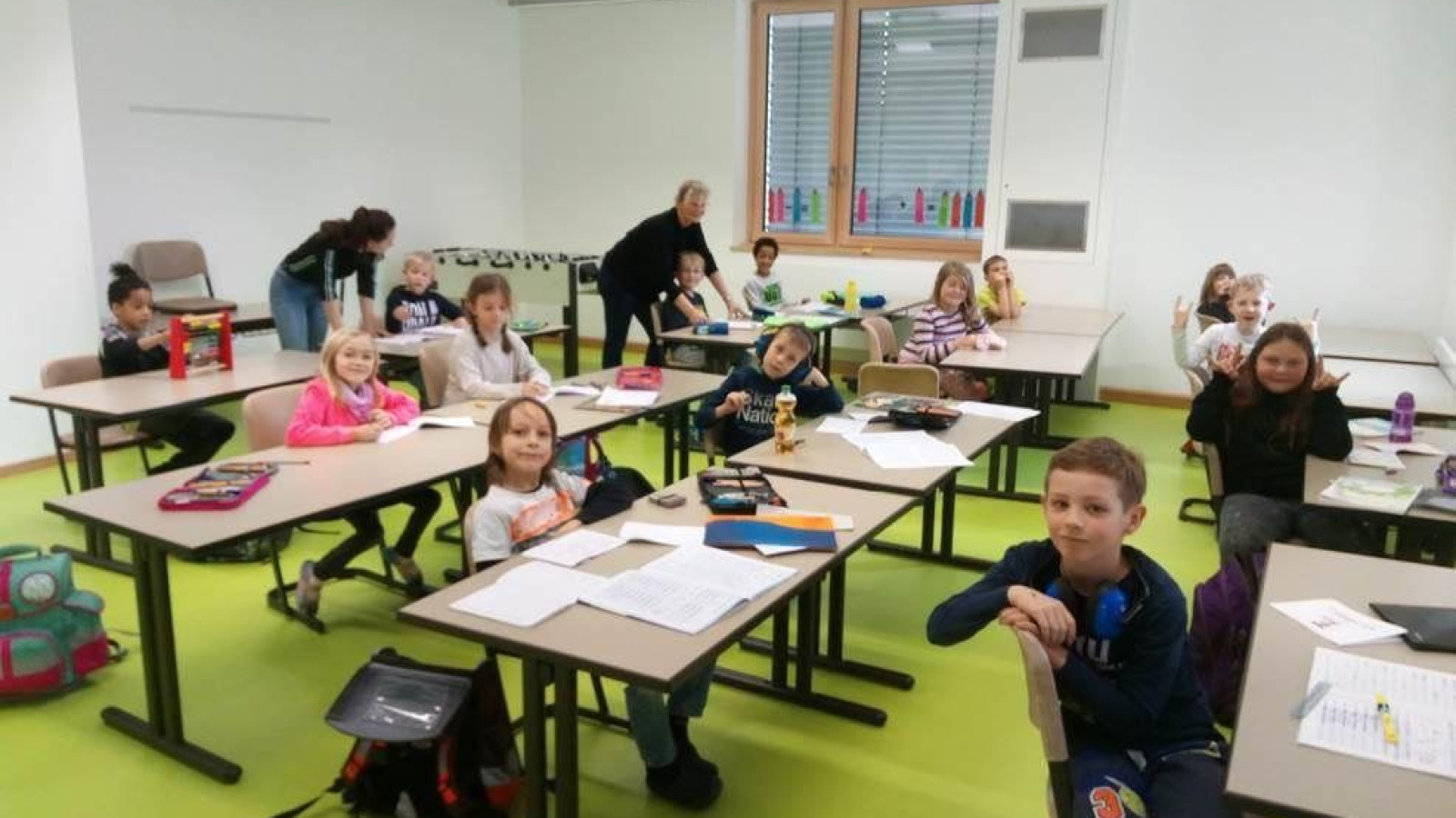 Mehrere Kinder und eine Lehrerin in einem modernen Klassenzimmer. Die Kinder sitzen an Tischen.