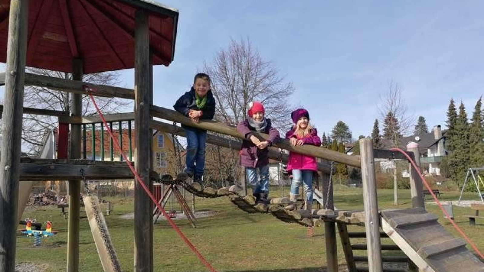 Vier Kinder auf einem Holzspielplatzgerät, sonniger Tag, leere Wiese im Hintergrund.