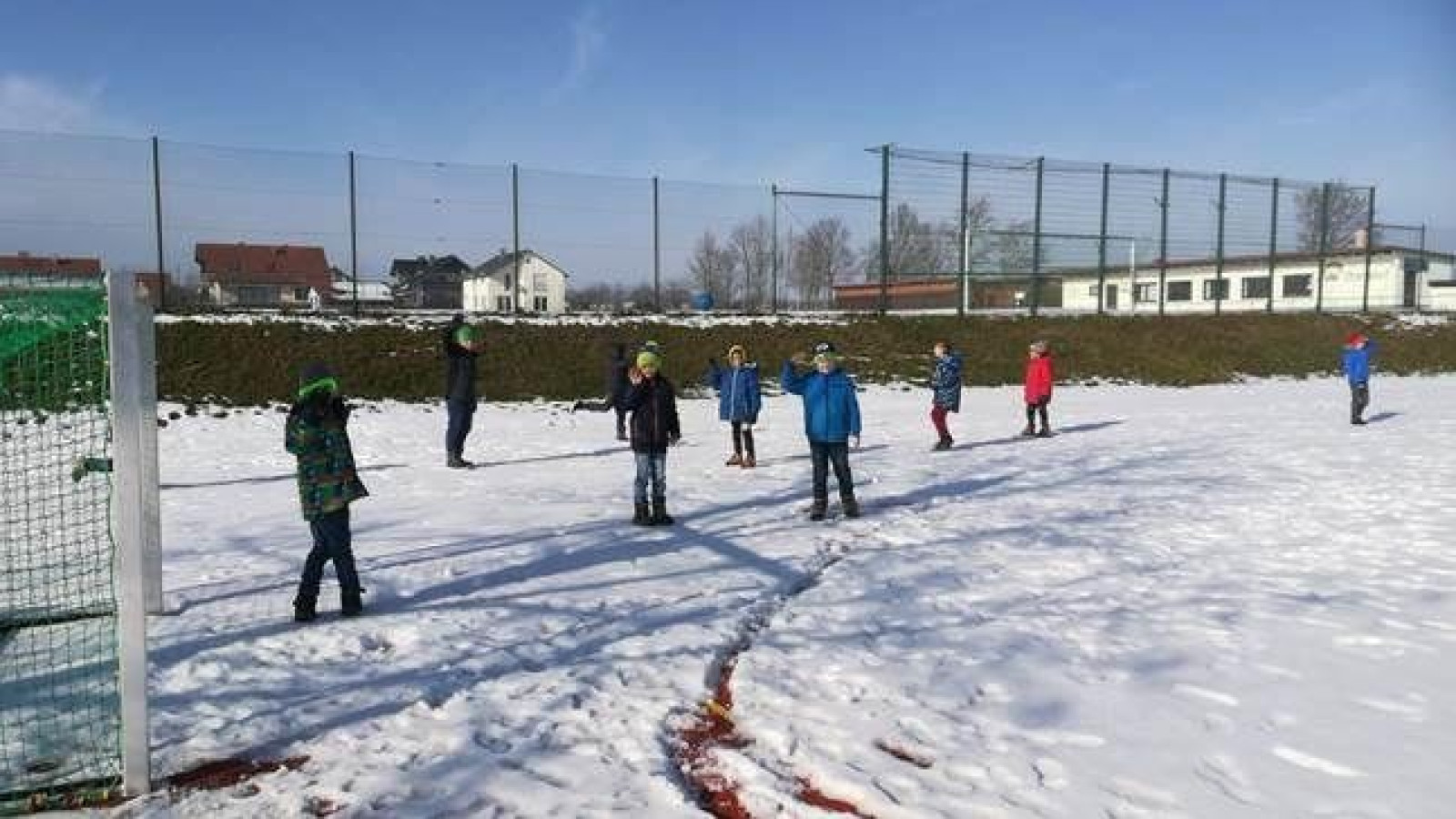 Kinder spielen im Schnee auf einem Fußballfeld, während ein Erwachsener am Spielfeldrand steht.