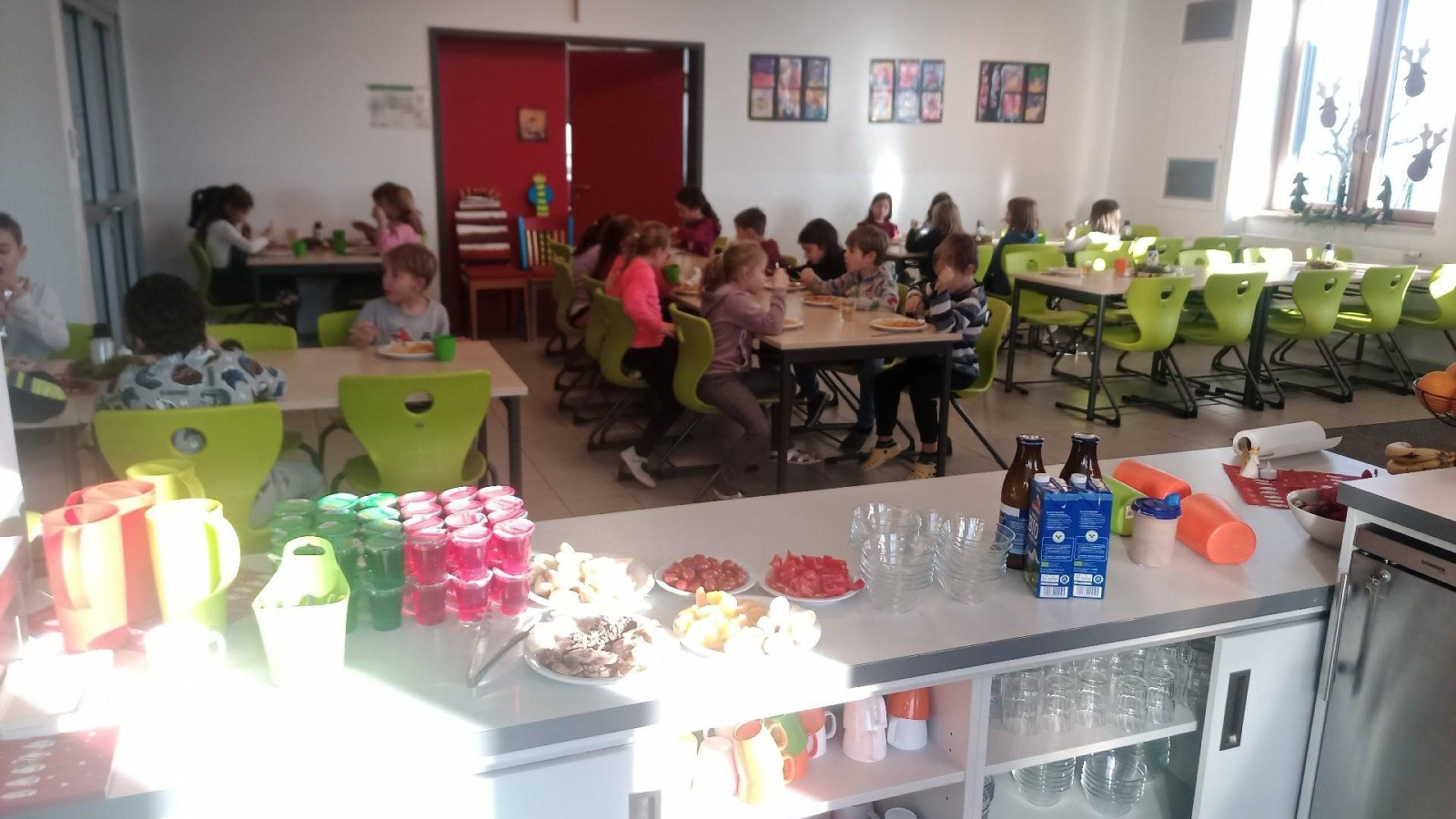 Kinder sitzen an Tischen in einer Schulkantine, mit Getränken und Snacks im Vordergrund auf einem Tresen.