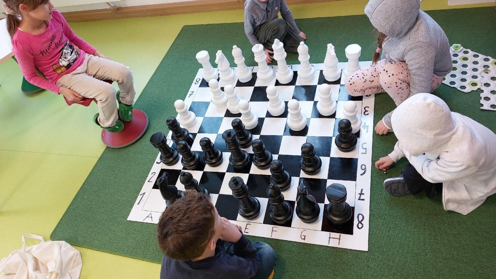 Kinder spielen Schach, sitzen auf grünem Boden rund um ein großes Schachbrett.