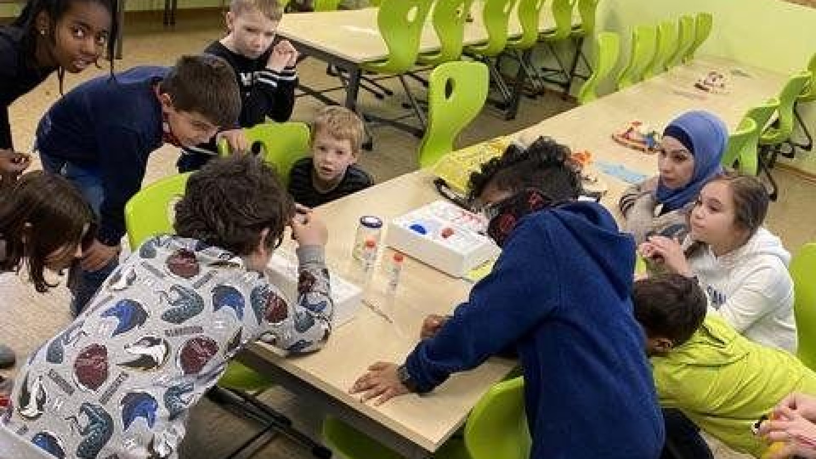 Kinder stehen in einem Klassenraum um einen Tisch, auf dem ein Wissenschaftsexperiment stattfindet.