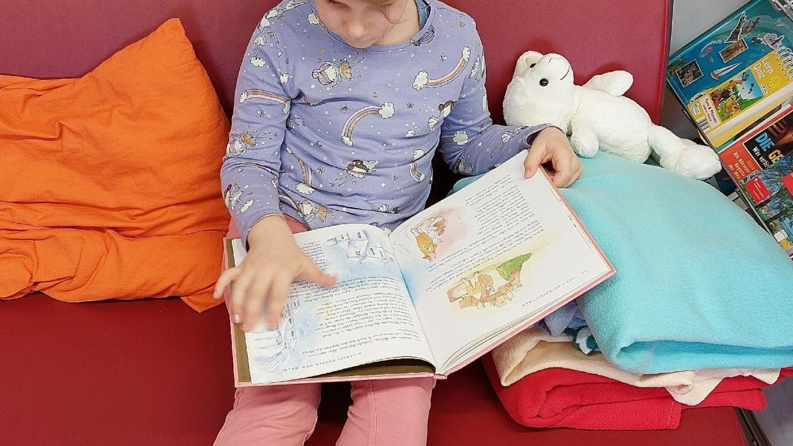 Ein Mädchen sitzt auf einer roten Couch mit einem Buch in den Händen und einem Stofftier neben sich.