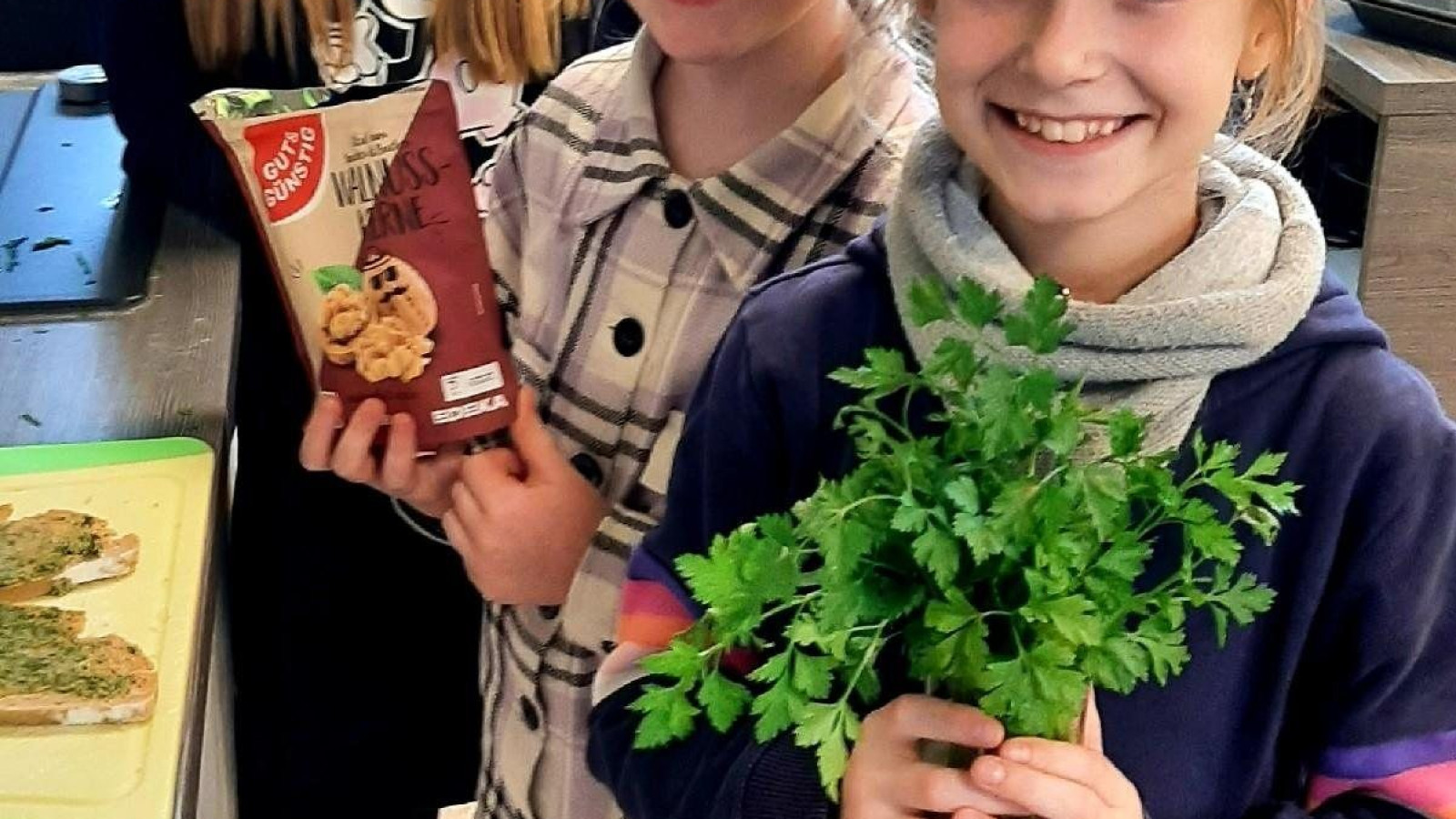 Drei Kinder in einer Küche, eines hält Kekse, das andere frische Kräuter.