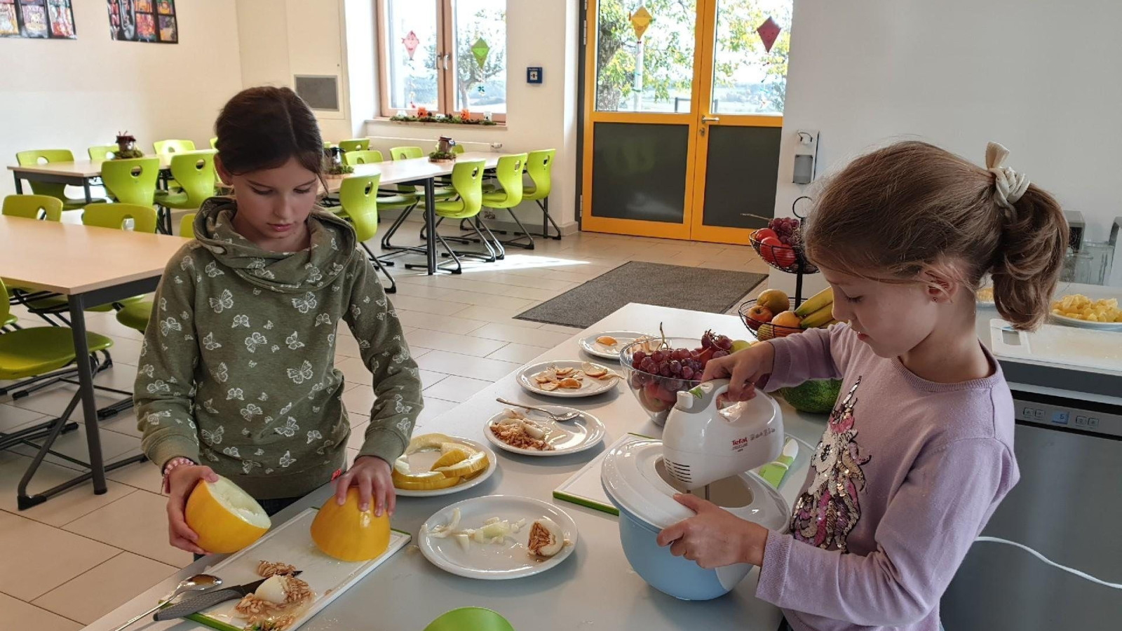 Zwei Kinder arbeiten in einer Schulküche, mit Lebensmitteln auf dem Tisch und leuchtend grünen Stühlen im Hintergrund.