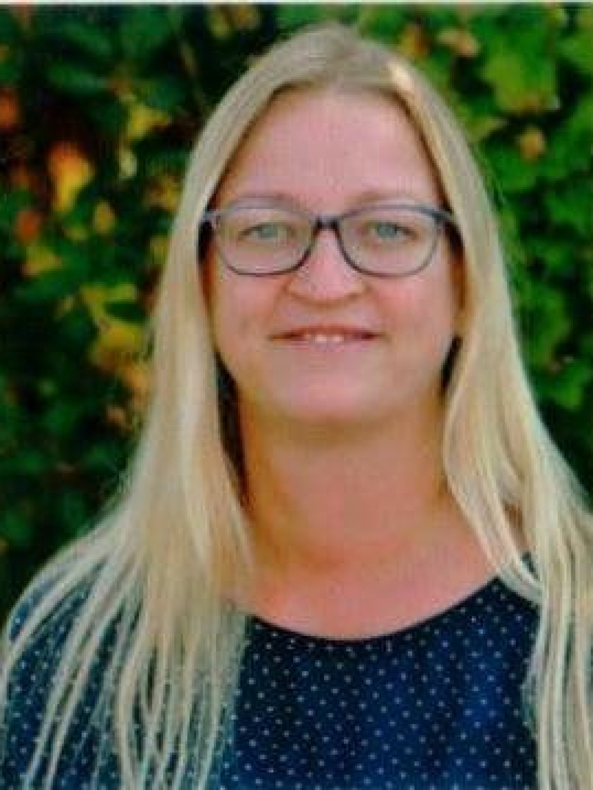 Frau mit langen blonden Haaren und Brille vor grünen Blättern.