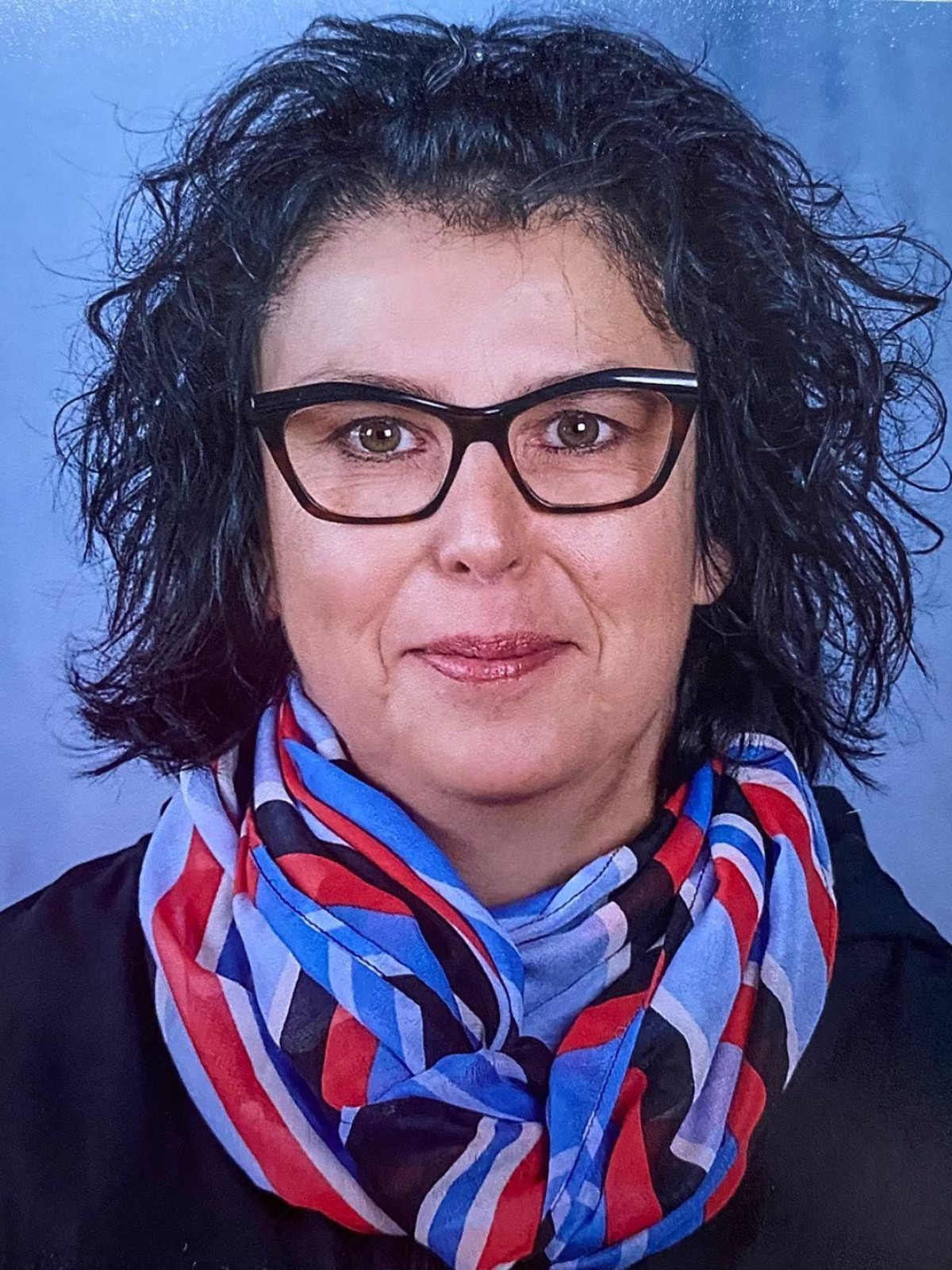 Porträt einer Frau mit schwarzem, lockigem Haar, Brille und einem bunten Schal vor blauem Hintergrund.