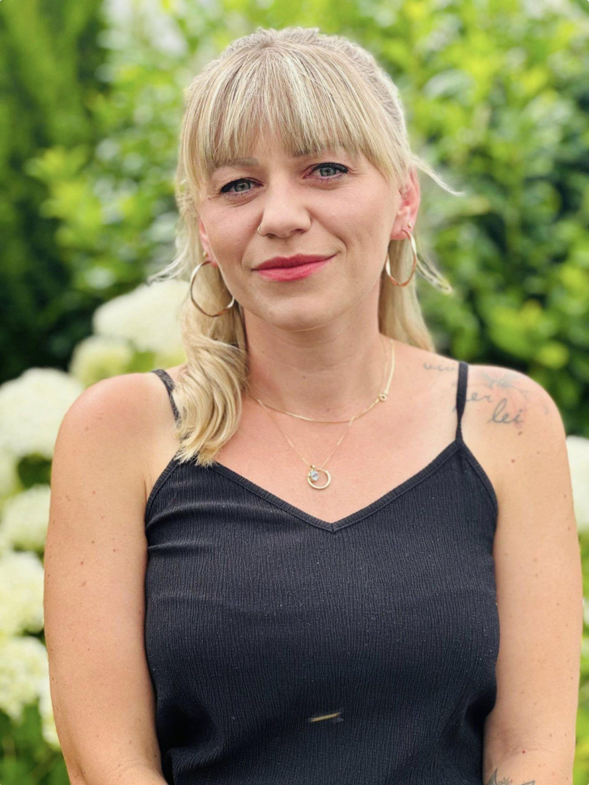 Blonde Frau in einem schwarzen Trägeroberteil vor einem Hintergrund aus grünen Pflanzen und weißen Hortensien.
