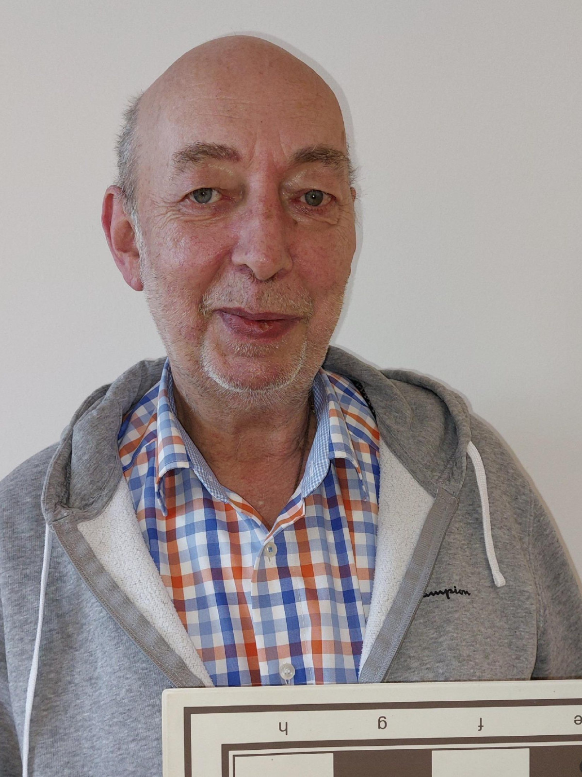 Ein Mann hält ein Schachbrett und trägt ein kariertes Hemd und einen grauen Hoodie.