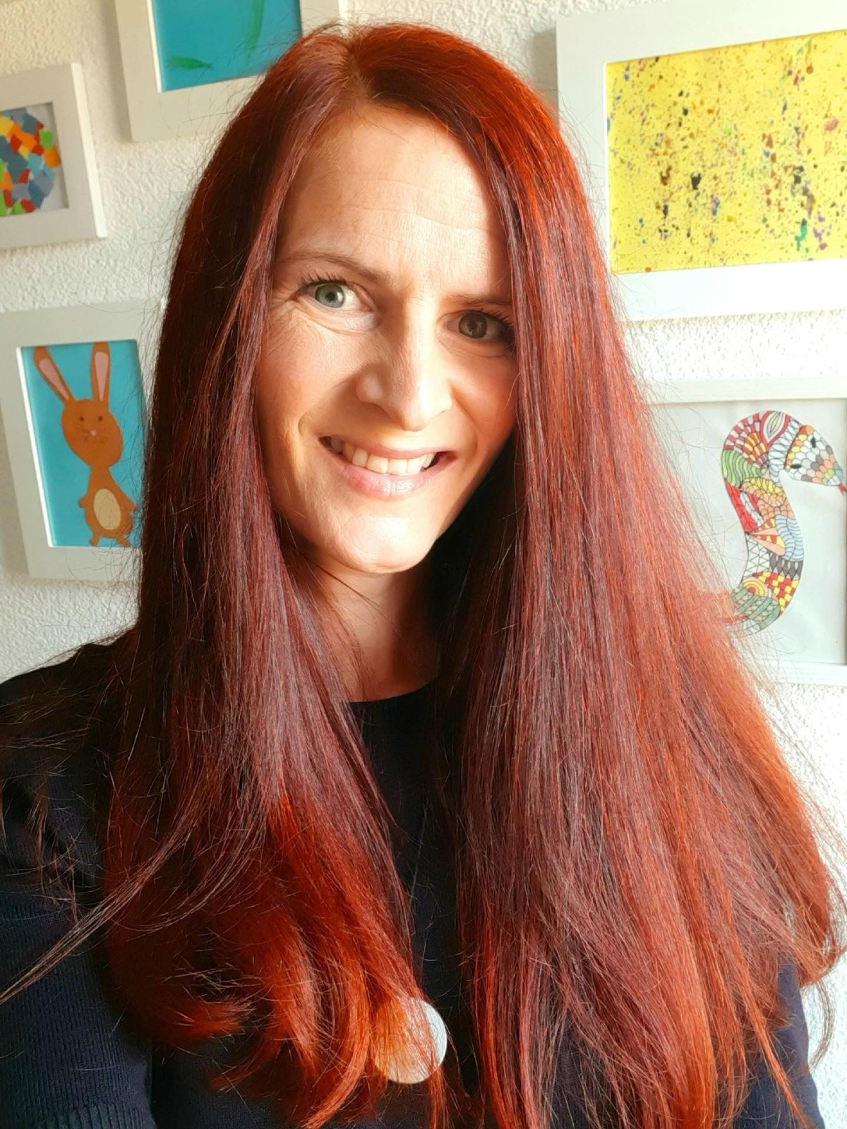 Frau mit langen roten Haaren lächelt, im Hintergrund sind bunt gerahmte Bilder an der Wand.
