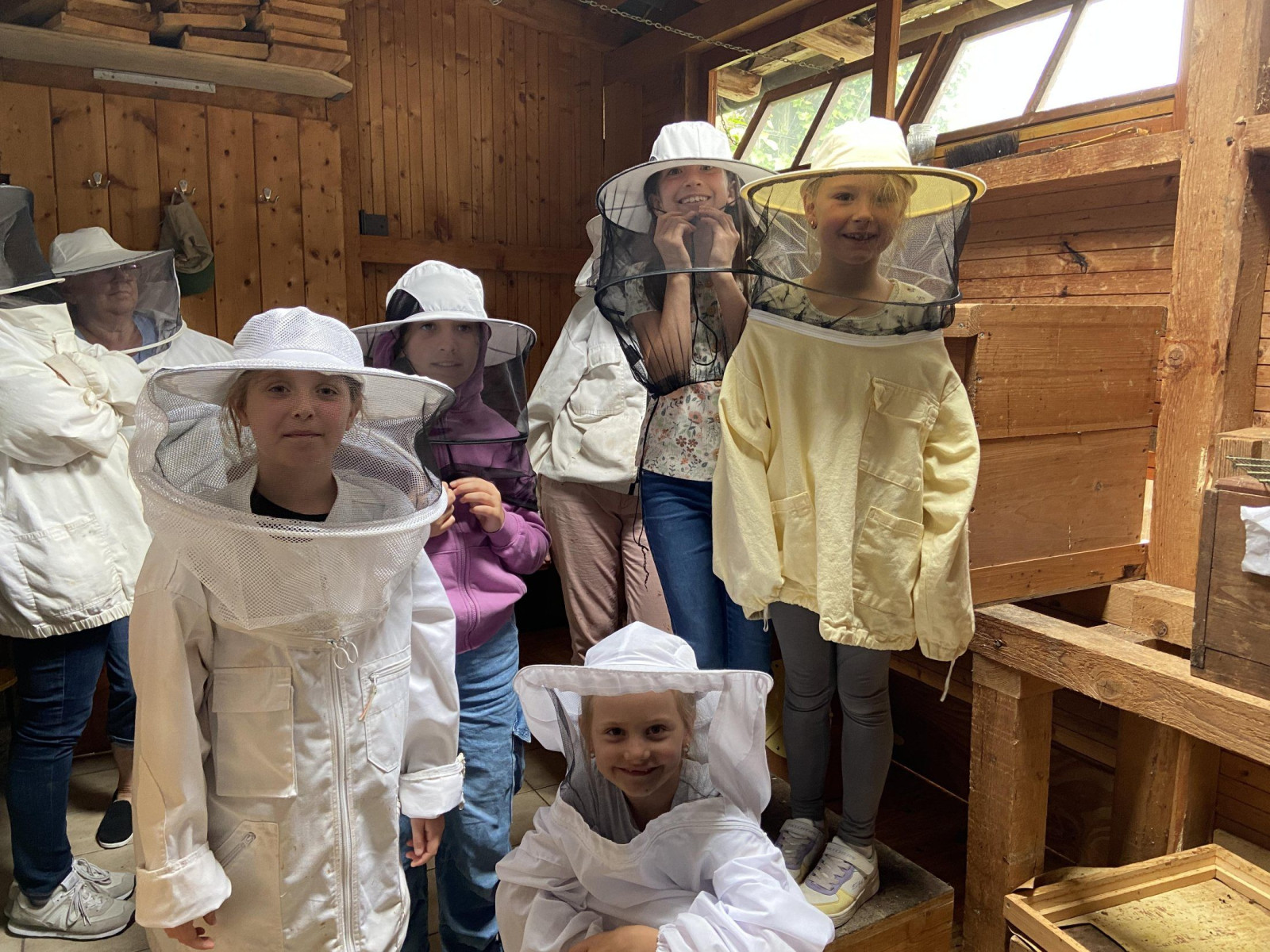 Kinder in Imkeranzügen stehen in einem hölzernen Bienenhaus mit offenen Fenstern und Holzkisten.