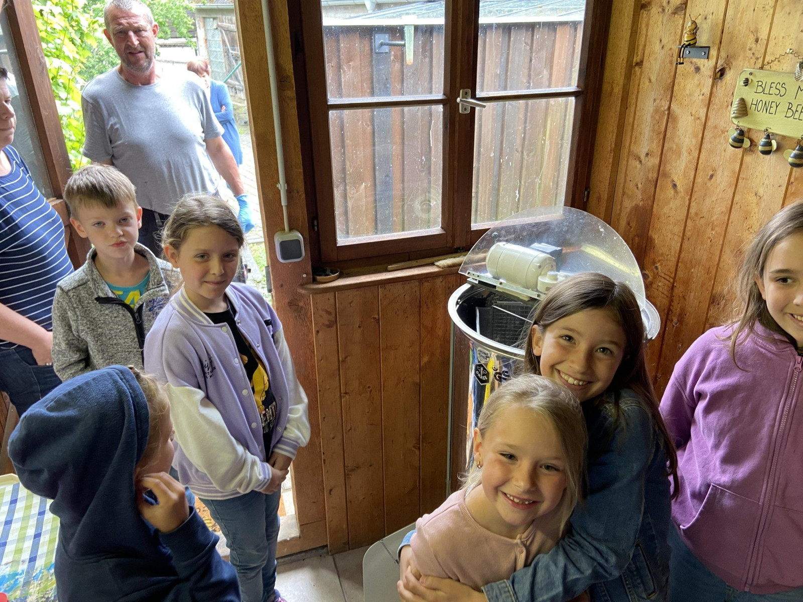 Kinder stehen um eine Honigschleuder in einer Holzhütte und lächeln in die Kamera.