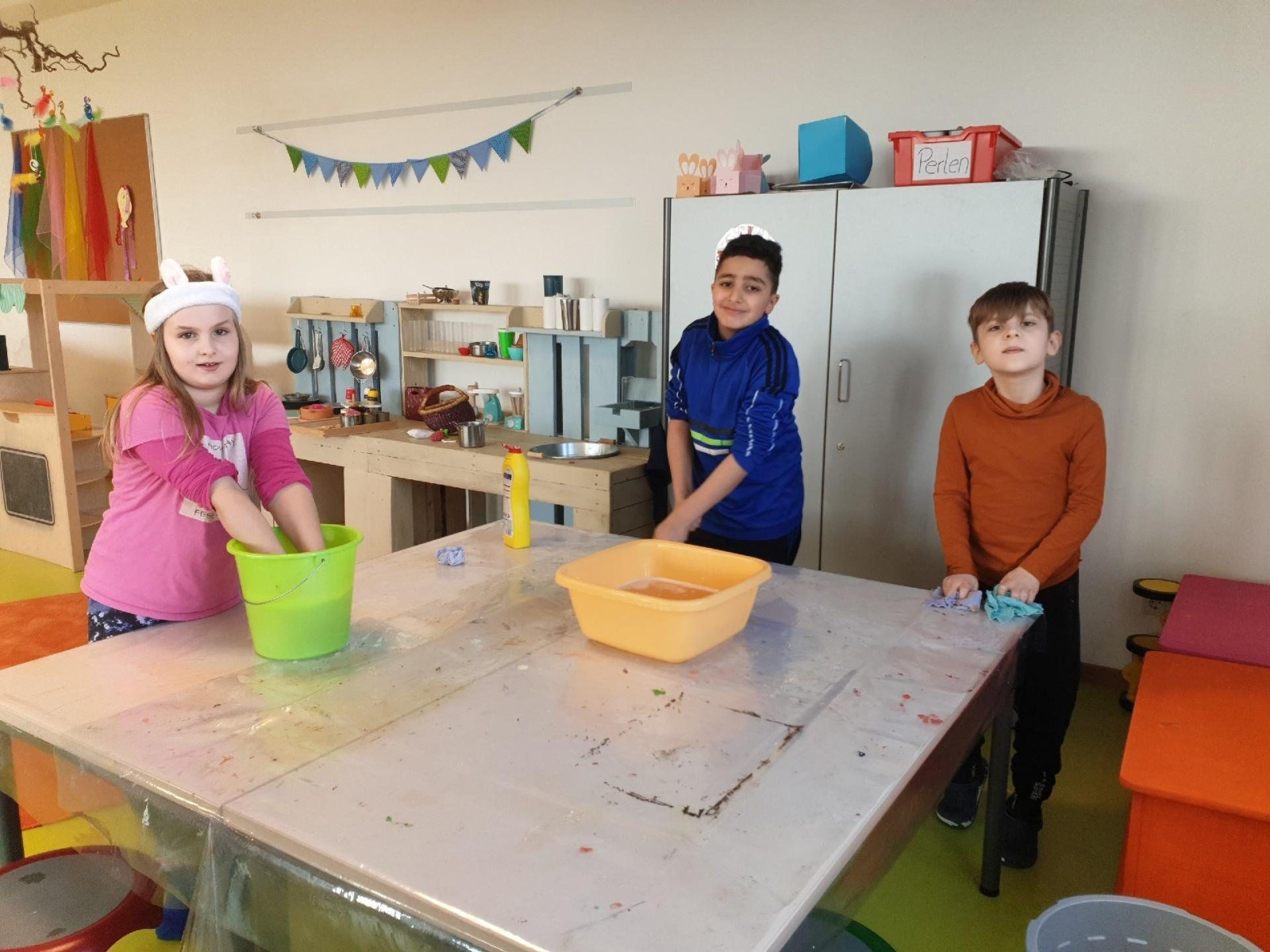 Drei Kinder spielen in einem Klassenzimmer mit Eimern und Lappen auf einem Tisch.