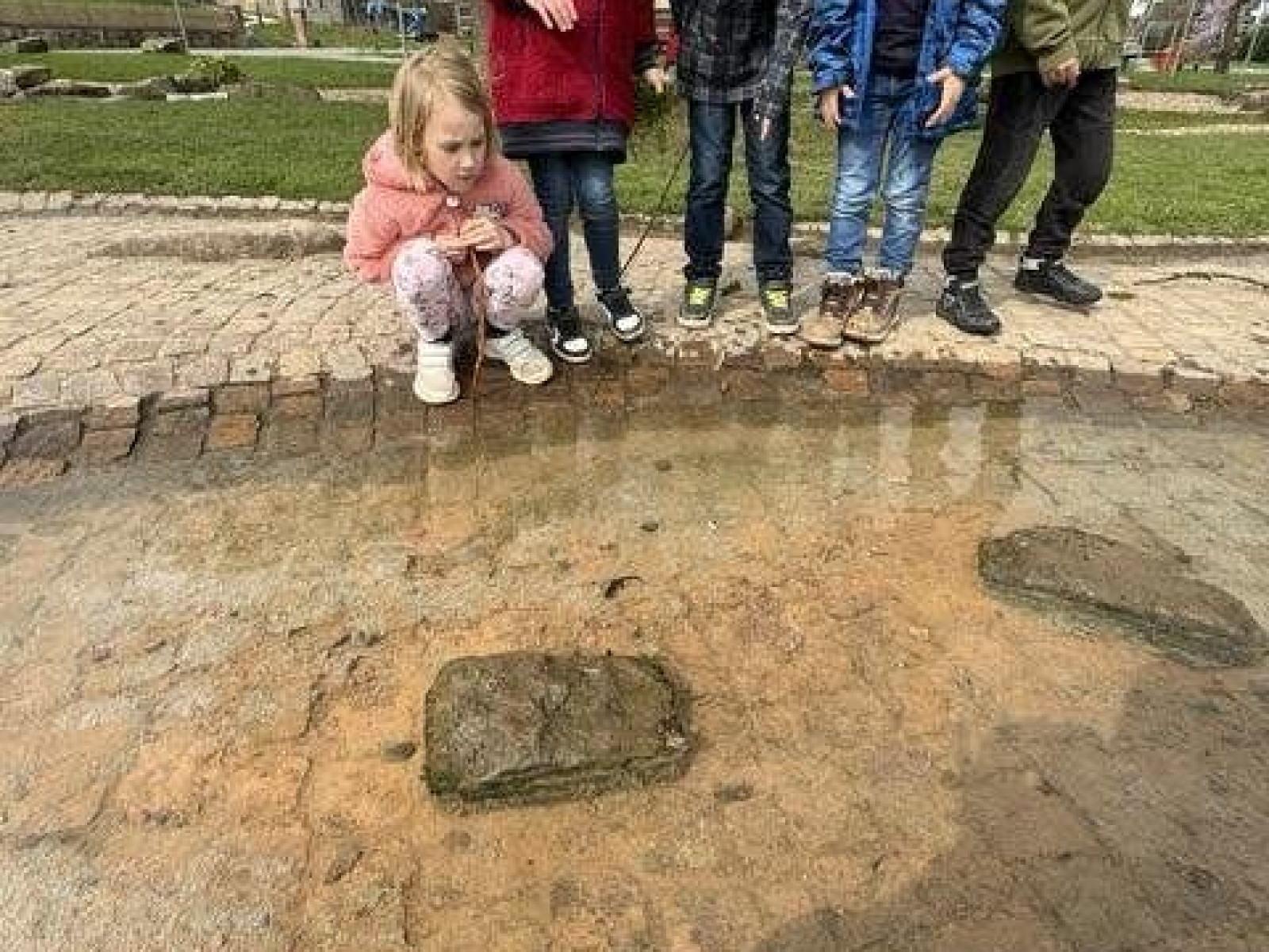 Fünf Kinder schauen auf ein flaches Wasserbecken auf einem Gehweg im Freien.