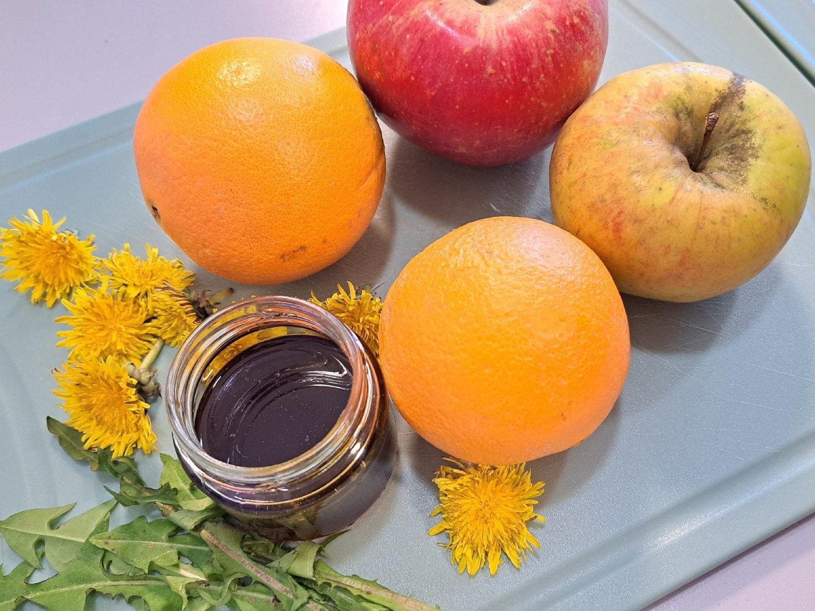 Ein Glas Honig, zwei Orangen, zwei Äpfel und Löwenzahnblüten auf einem Schneidebrett.