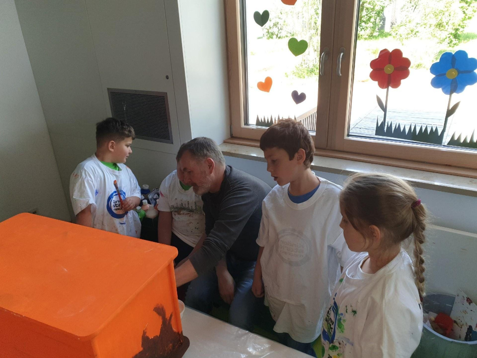 Kinder in weißen T-Shirts schauen einem Mann bei einer Arbeit zu, Fenster mit Blumen dekoriert.