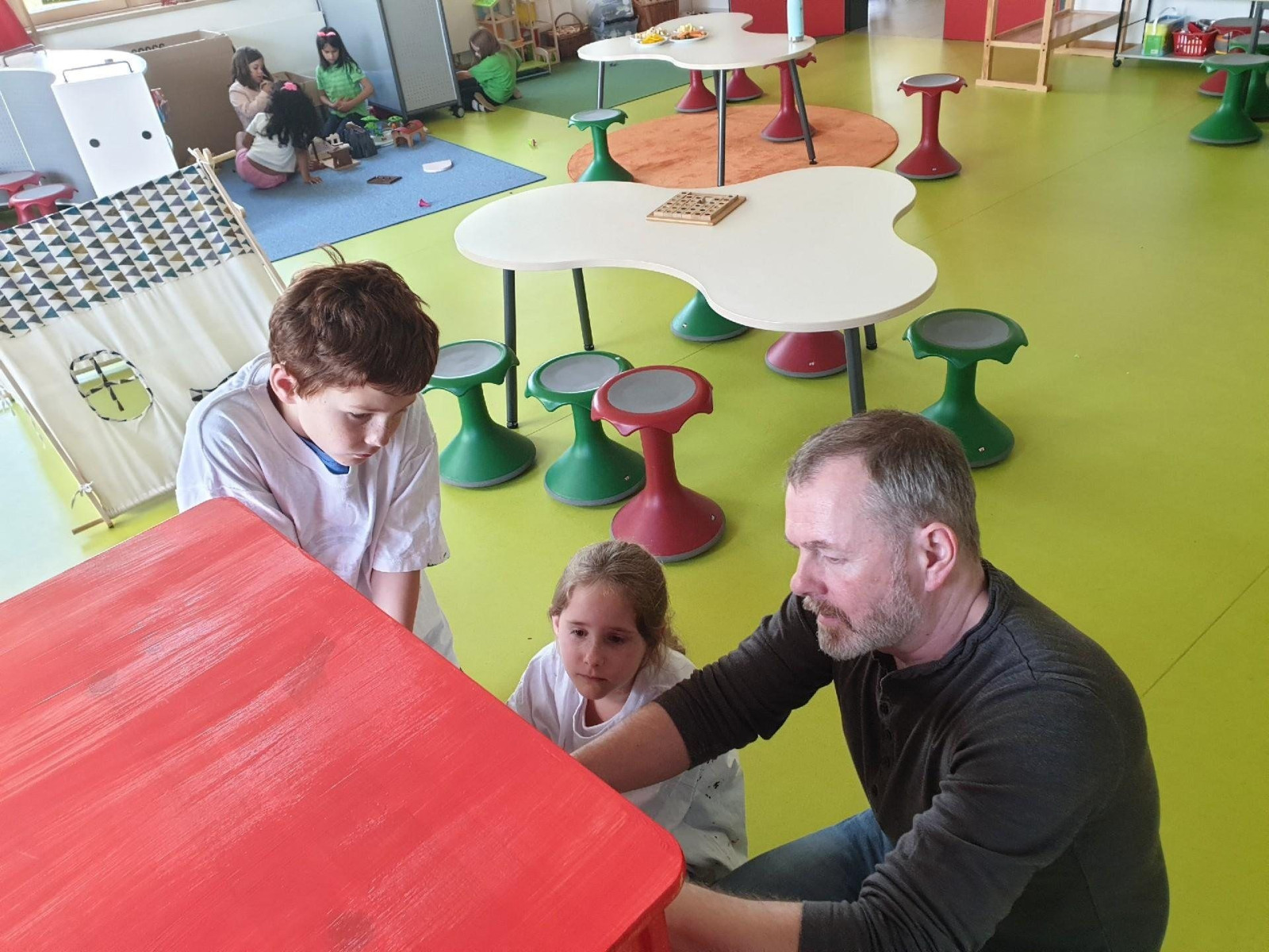 Mann und zwei Kinder bemalen ein Möbelstück in einem bunten Kita-Raum.