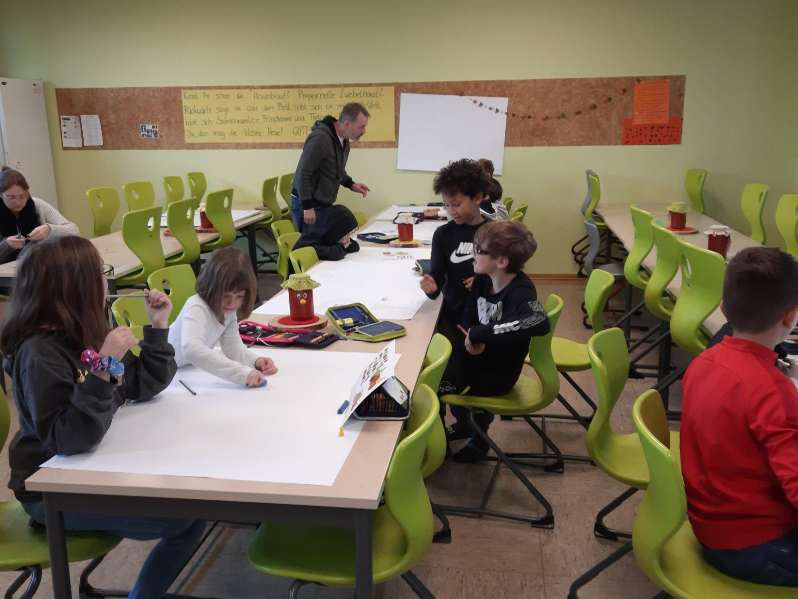 Kinder in einem Klassenzimmer, die an einem Tisch basteln, umgeben von grünen Stühlen.