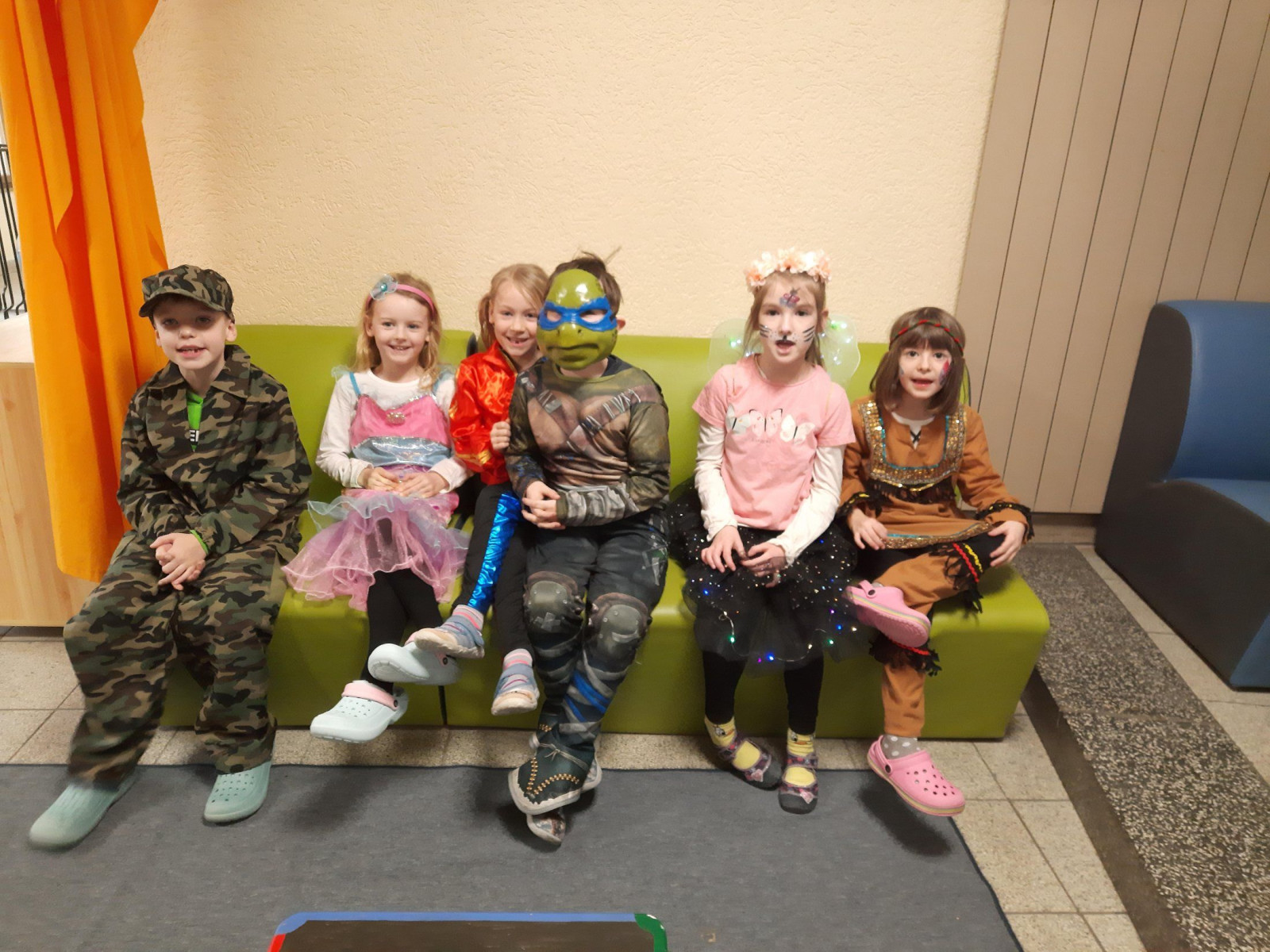 Sechs Kinder in farbenfrohen Kostümen sitzen auf einer grünen Bank und lächeln.