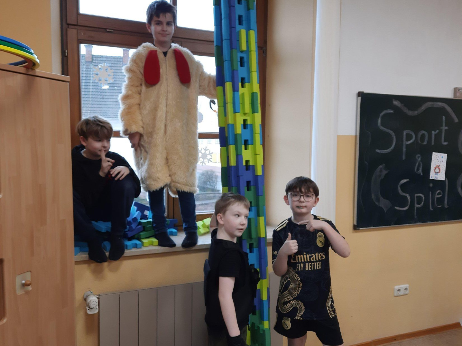 Vier Jungen präsentieren einen hohen Turm aus bunten Schaumstoffblöcken in einem Klassenzimmer.