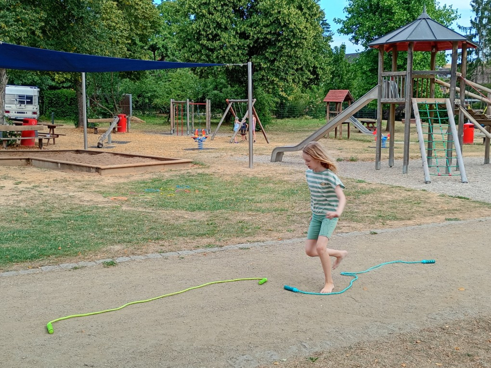 Kind springt über ein Springseil auf einem Spielplatz mit Rutschen und Klettergeräten.