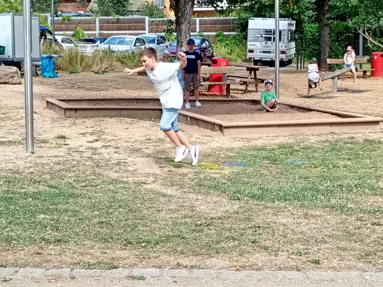 Junge springt auf einem Spielplatz, andere Kinder im Hintergrund.