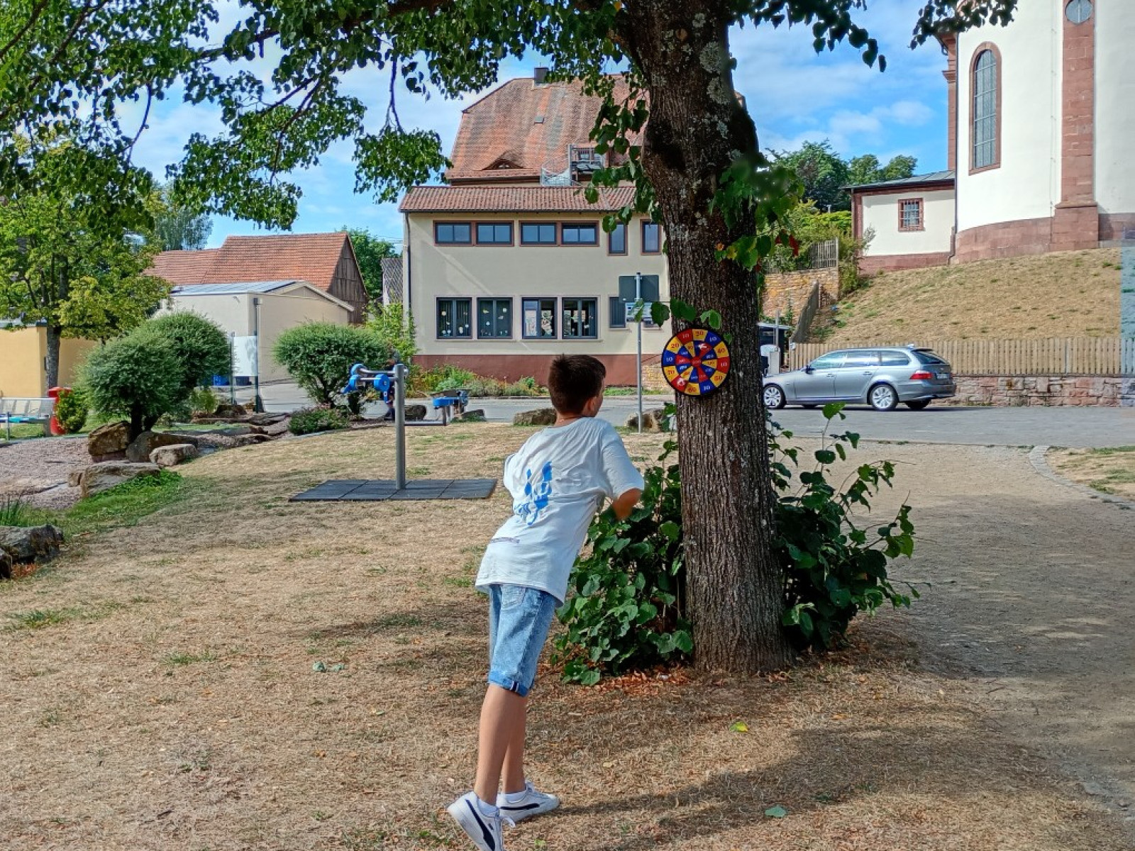 Junge wirft Dartpfeile auf eine Zielscheibe an einem Baum, mit Häusern und Kirche im Hintergrund.