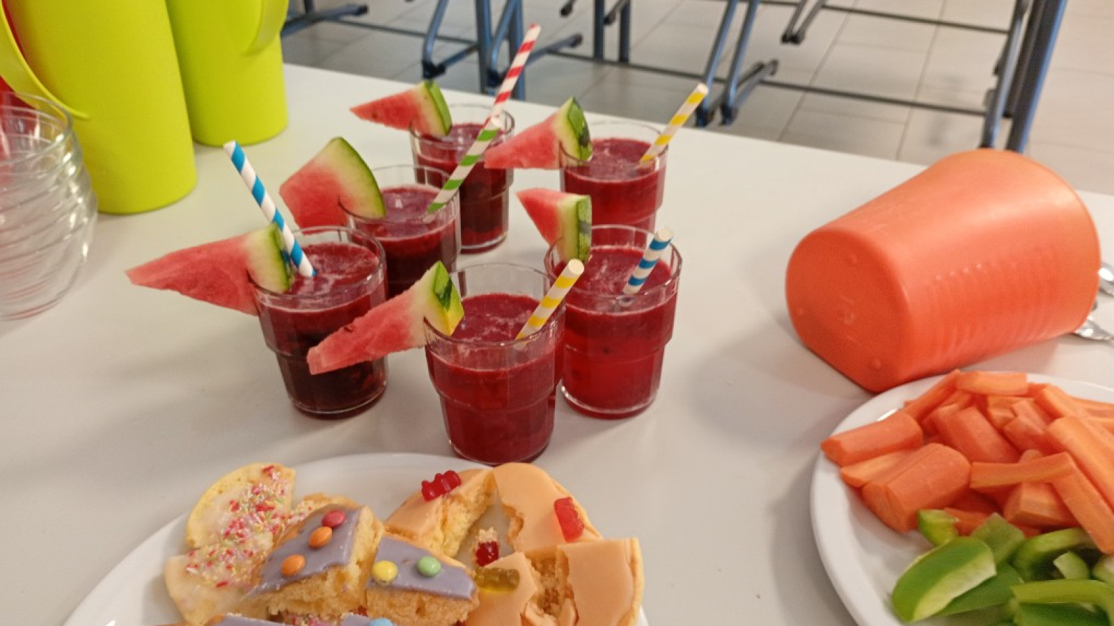 Sechs Gläser mit rotem Getränk und Wassermelonen-Dekoration, Kekse und Gemüsestücke auf einem Tisch.