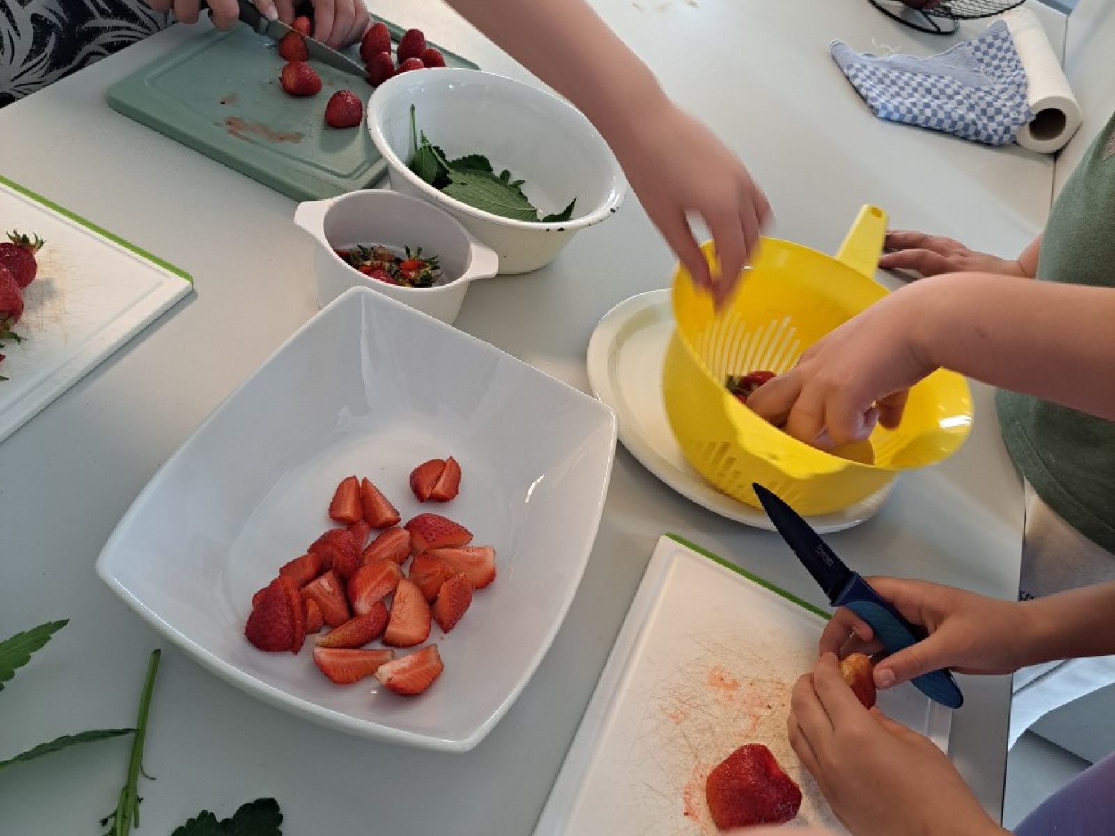 Personen schneiden Erdbeeren auf einem Tisch. Eine gelbe Zitronenpresse steht daneben.
