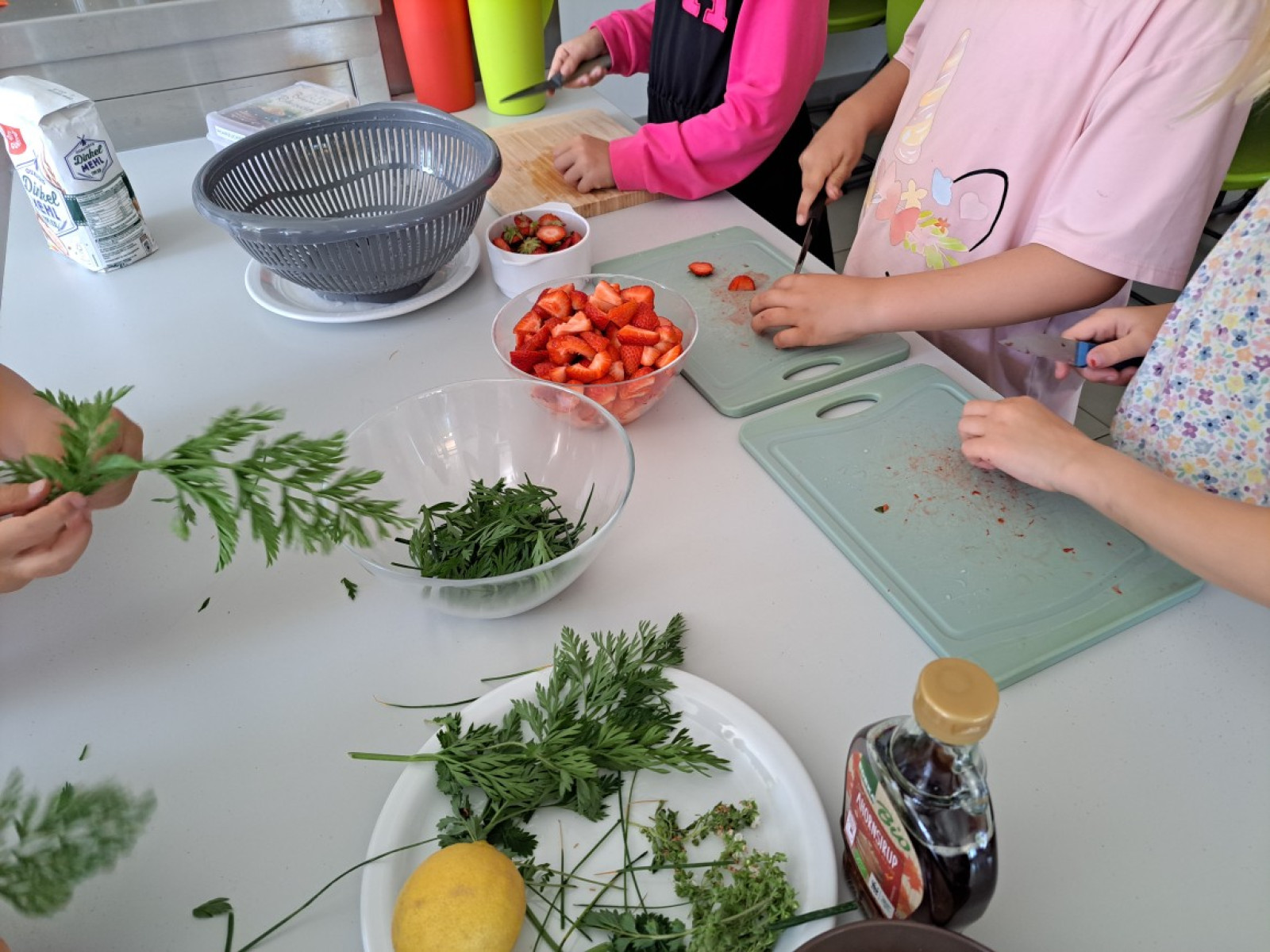 Kinderhände schneiden Erdbeeren und bereiten Kräuter an einem Tisch mit Küchenutensilien vor.
