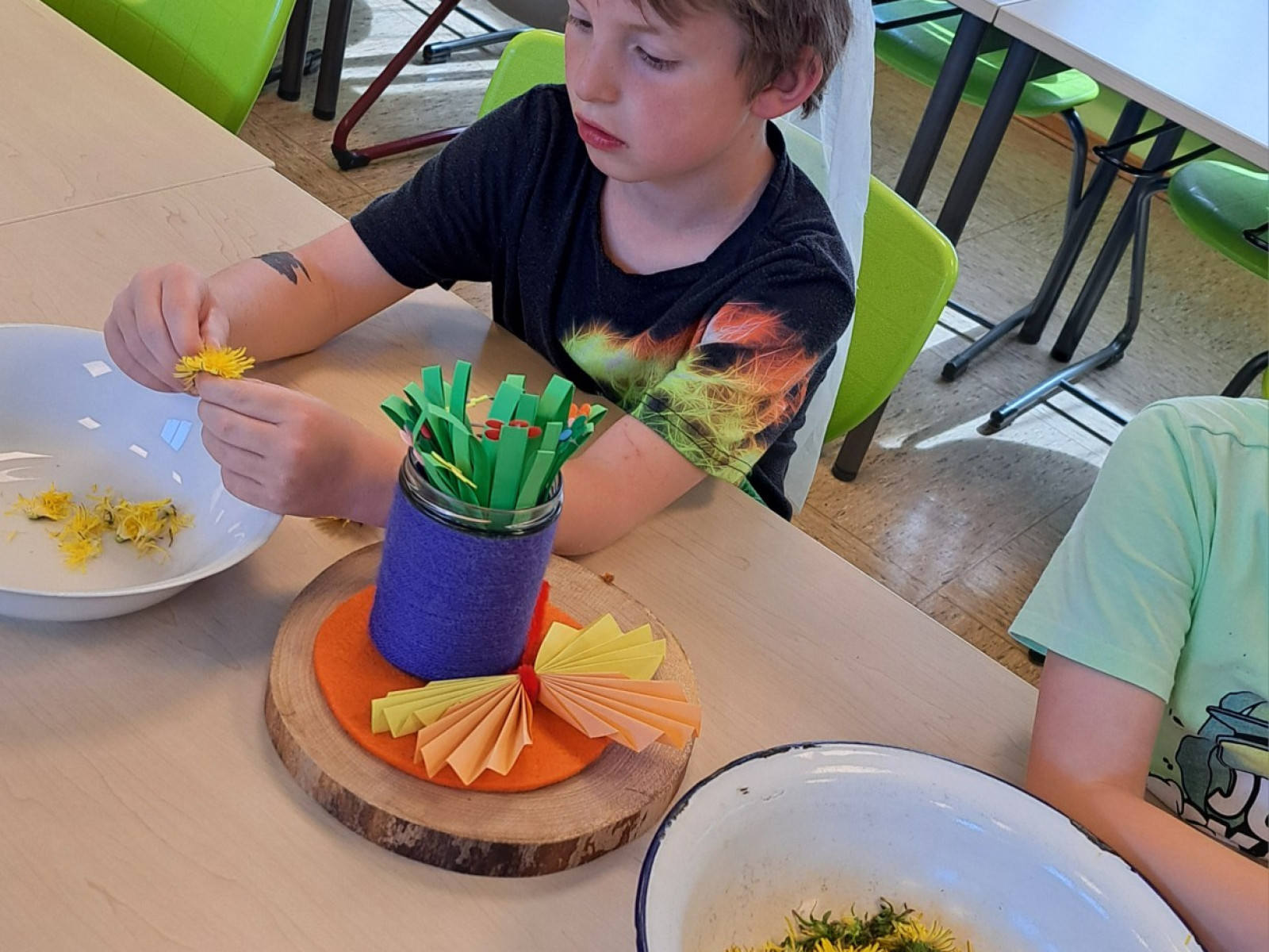 Junge mit Krone bastelt mit Löwenzahnblüten und Papierstreifen in einer Schulklasse.
