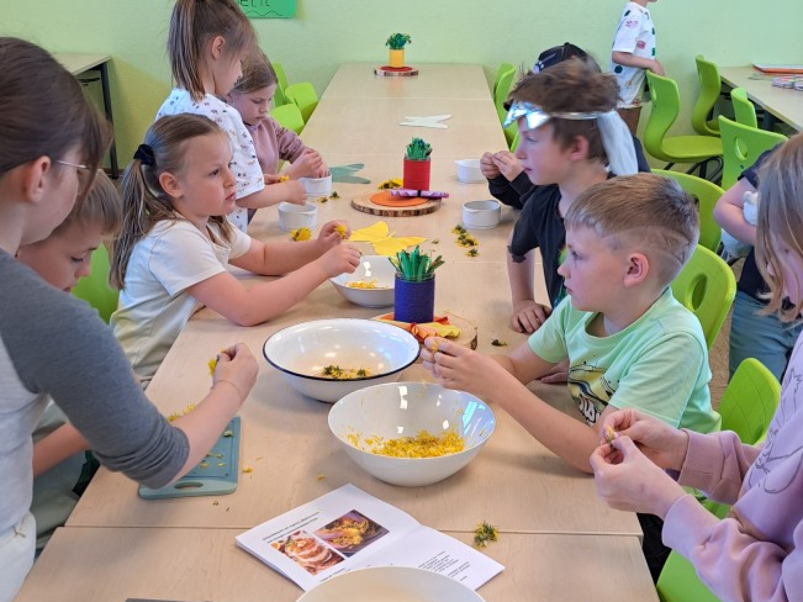 Kinder sitzen an einem langen Tisch und schneiden Obst in einem hellgrünen Klassenzimmer.