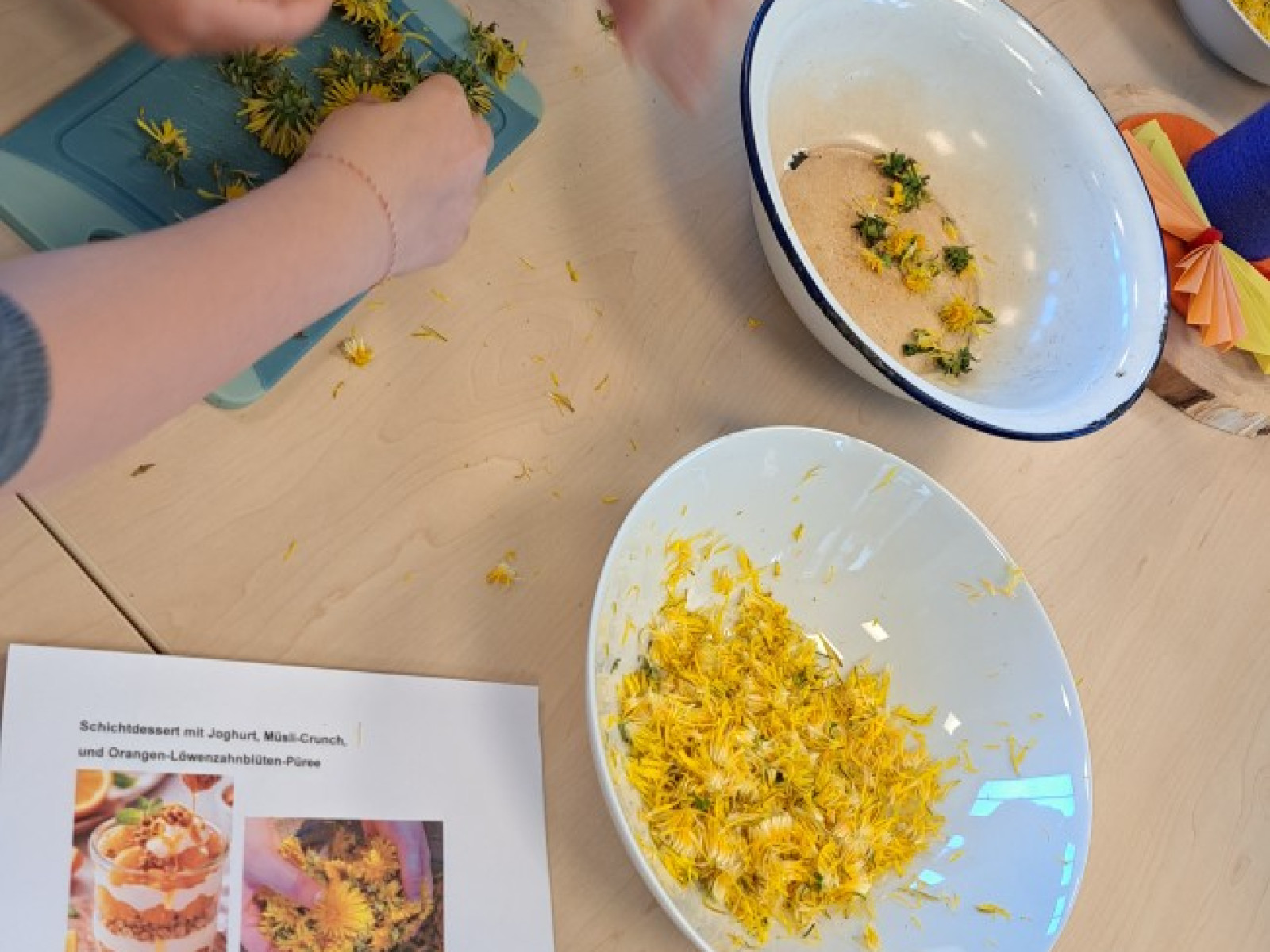 Kinder verarbeiten Löwenzahnblüten an einem Tisch, umgeben von Schalen und einem Rezeptblatt.